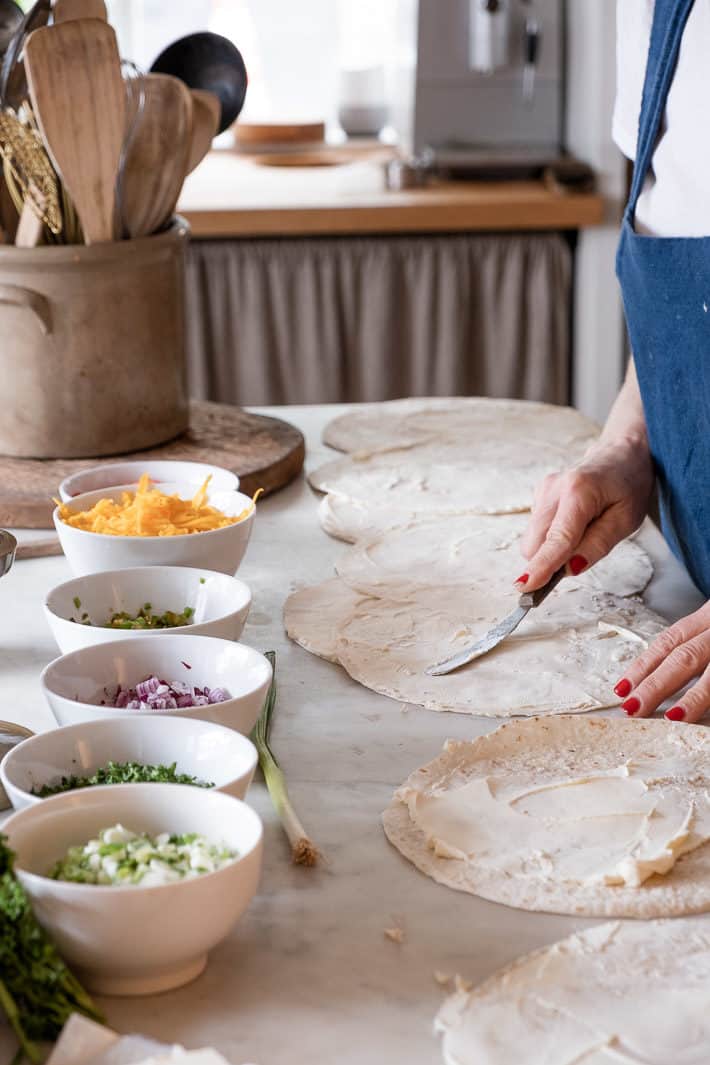 An assembly line of ingredients line a marble counter as a woman spreads cream cheese on tortillas.