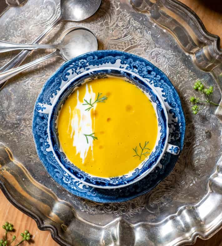 Overhead shot of orange, carrot soup garnished with swirls of sour cream, cilantro leaves and oil - in a blue and white transferware bowl on a silver tray.