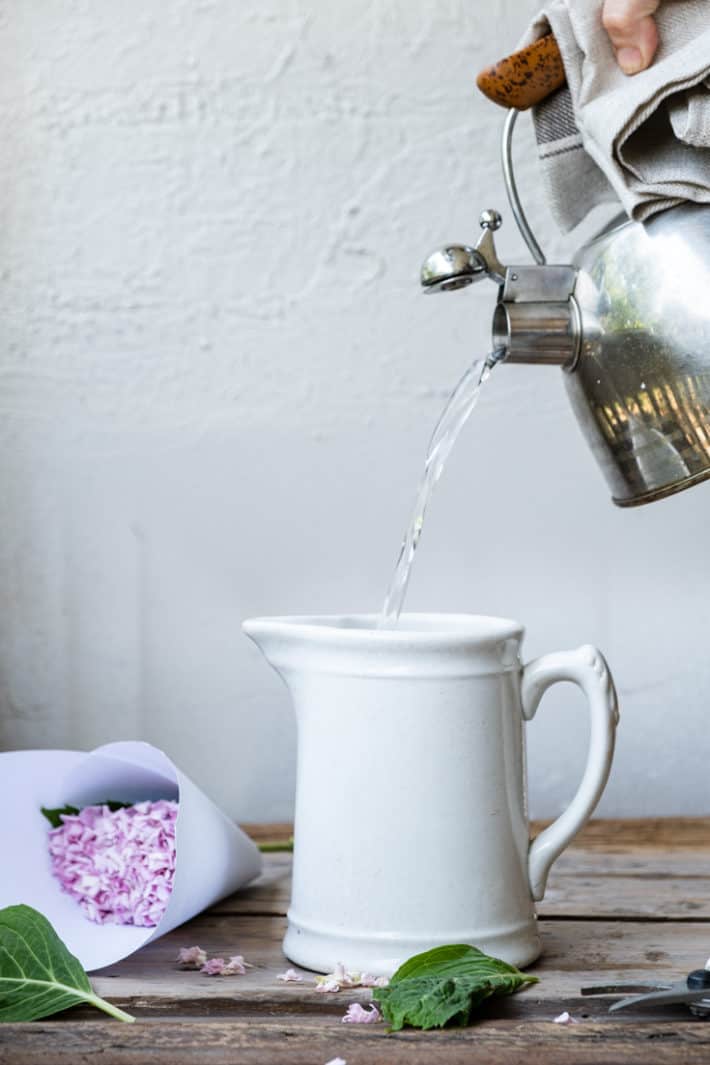 Hot water being poured from a kettle into an ironstone jug with a hydrangea laid to the side.