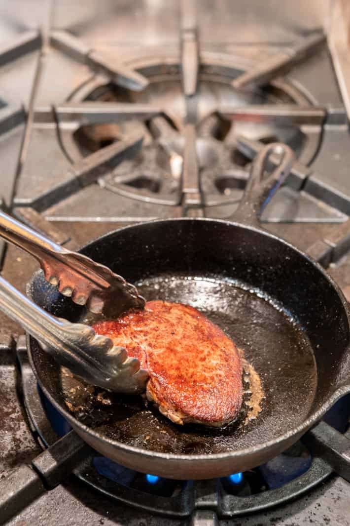 Flipping a cooking steak with tongs in a cast iron pan.