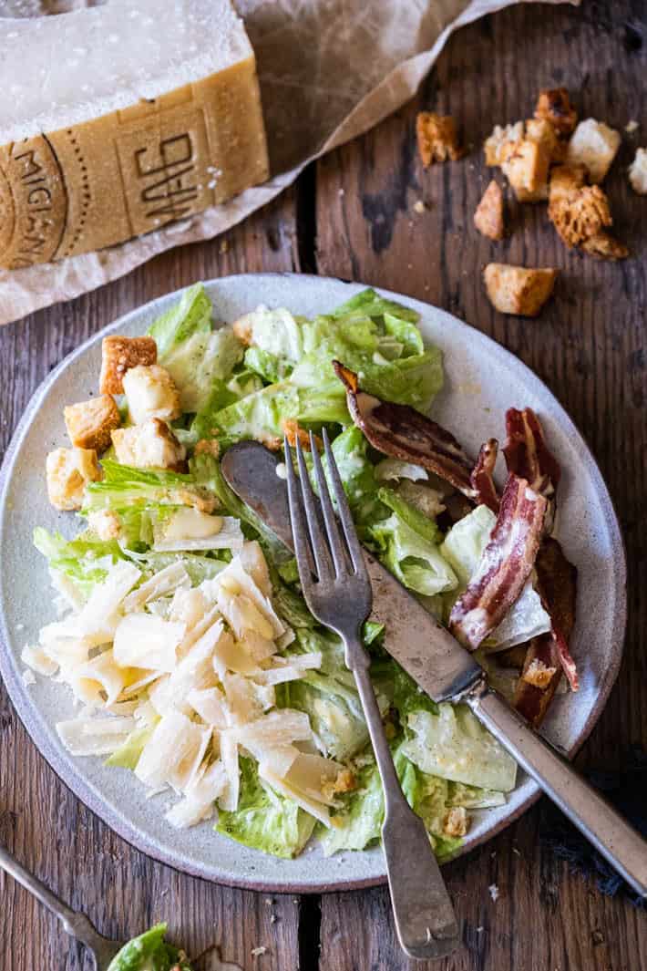 Caesar Salad on a handmade plate set on wood table with a wedge of parmesan cheese and croutons to the side.