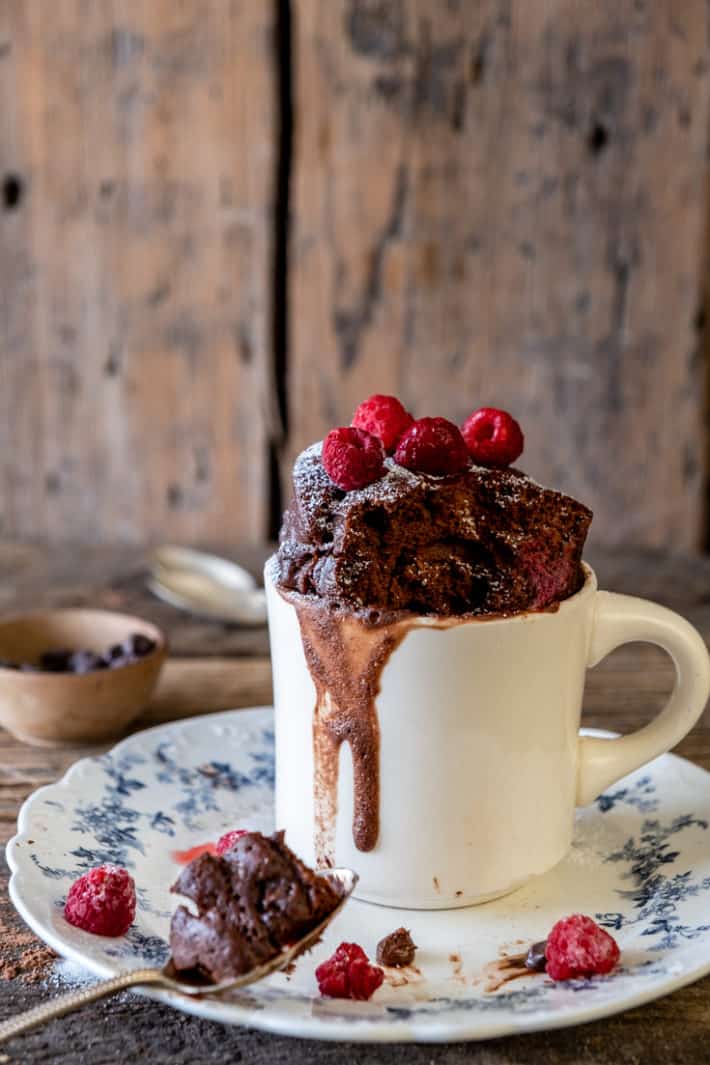 Cooked chocolate cake in a cup rising up from an ironstone mug topped with bright red raspberries and powdered sugar.