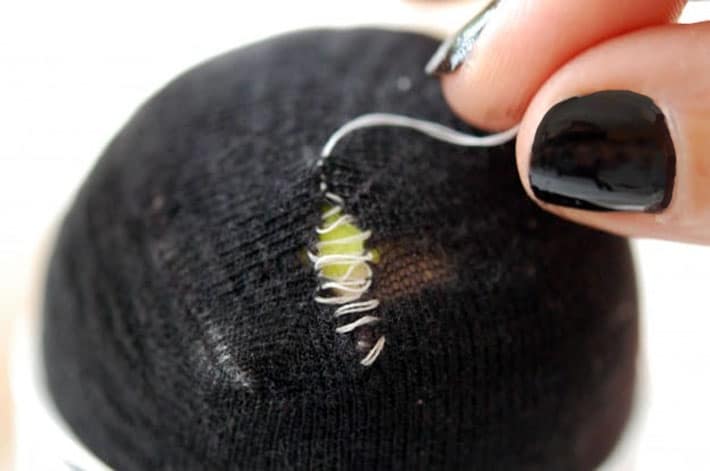 Black sock over yellow tennis ball being darned with white thread. Thread being pulled to tighten stitching.