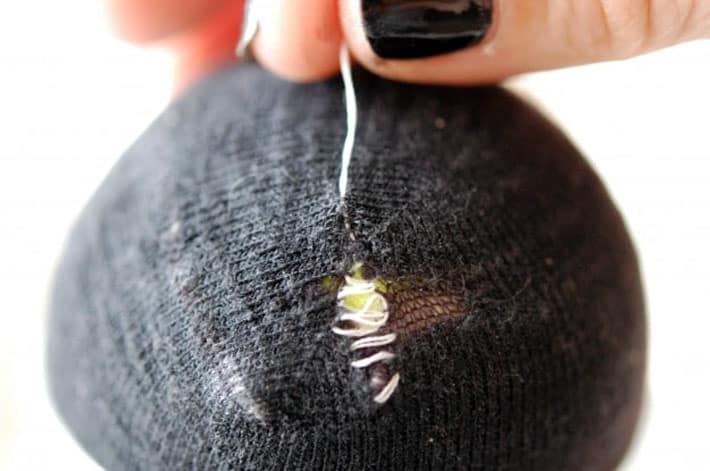 Black sock over yellow tennis ball being darned with white thread being pulled to tighten stitches.