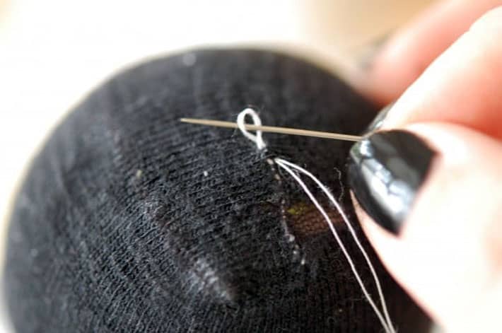 How to darn a sock. Black sock over yellow tennis ball being darned with white thread. Finishing mend by making a knot.