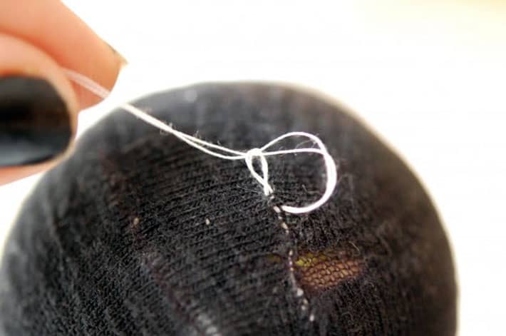 How to darn a sock. Black sock over yellow tennis ball being darned with white thread. Finishing mend by making a knot.