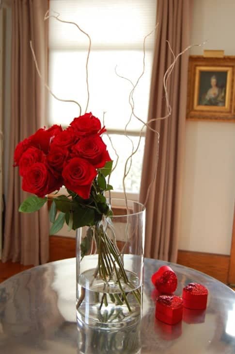 Contemporary arrangement of a dozen red roses tied together in a bunch, leaning to the side in a clear cylinder vase with a few white curly branches for interest.