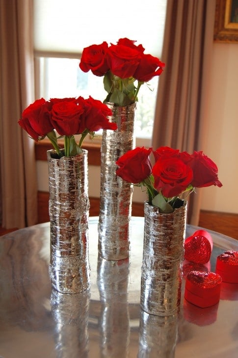 A dozen red roses divided among 3 narrow silver cylinder vases, with 4 roses in each vase.