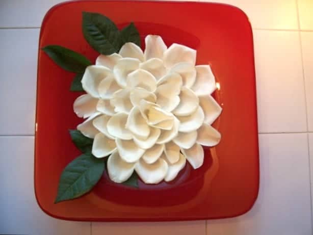 Red, shallow, glass bowl with what appears to be a huge white rose in the centre of it. It's actually individual petals floating on water to resemble a huge flower.