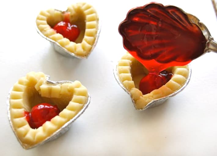 Antique spoon dabbing cherry pie filling into mini tarts on white counter.