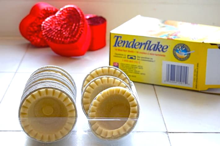 Frozen mini tart shells on a white tile counter with empty cardboard box and red sequin hearts.