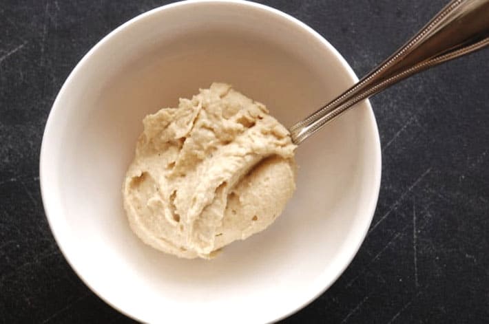Big spoon of hummus being dropped into a white bowl on a black countertop.
