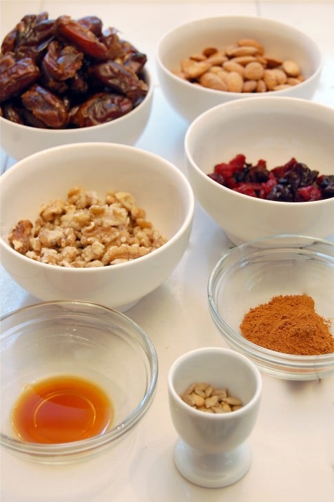 Various small bowls on a white counter filled with dates, almonds, walnuts, spices and cranberries.