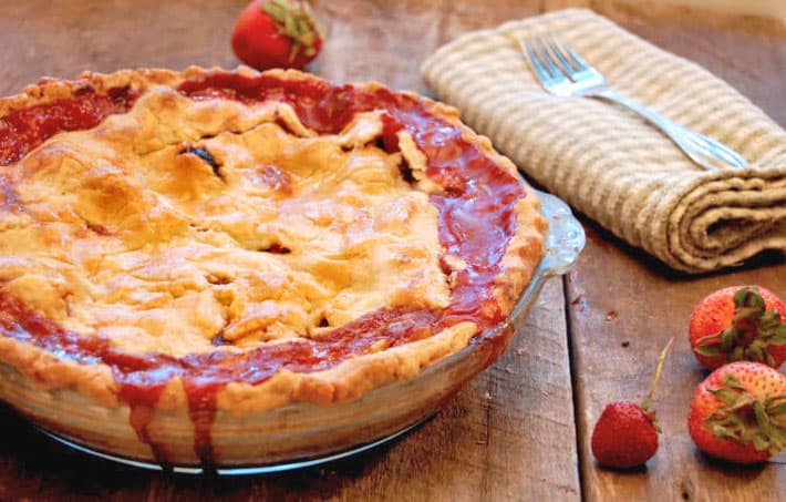 Strawberry rhubarb pie in a Pyrex dish on a wood table with strawberries scattered around.