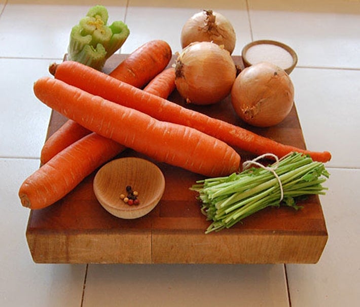 Carrots, celery, onions, parsley, salt and pepper on wood chopping block.