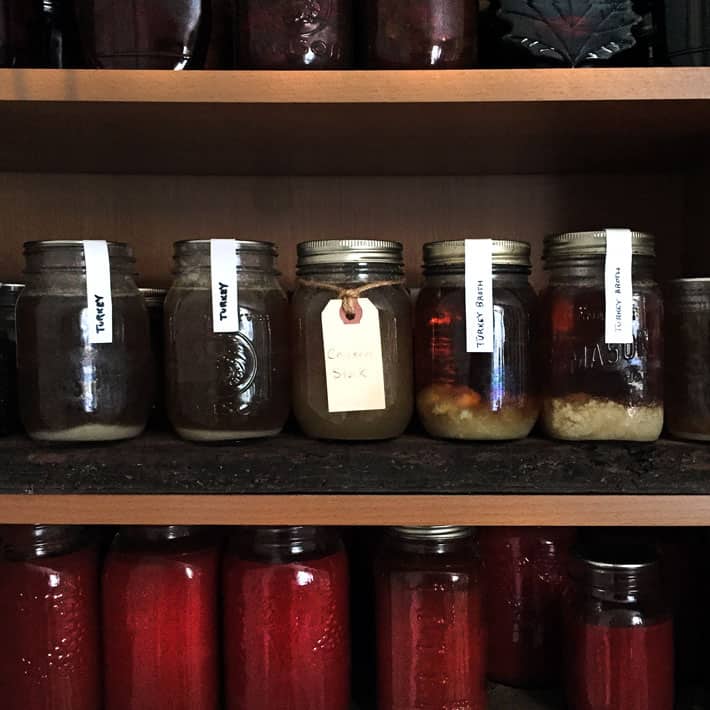 Wood pantry shelves filled with red tomato sauce on the bottom shelf, chicken broth on the middle shelf and maple syrup on the top shelf.