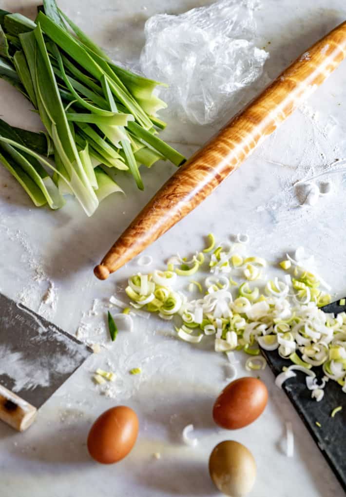 Flame birch rolling pin amidst dark green leek ends, sliced leek and brown and green eggs.