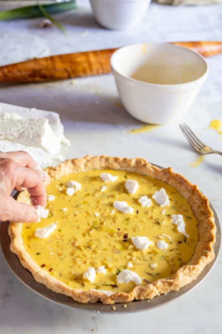 A leek quiche being topped with crumbled goat cheese with other ingredients in the background.