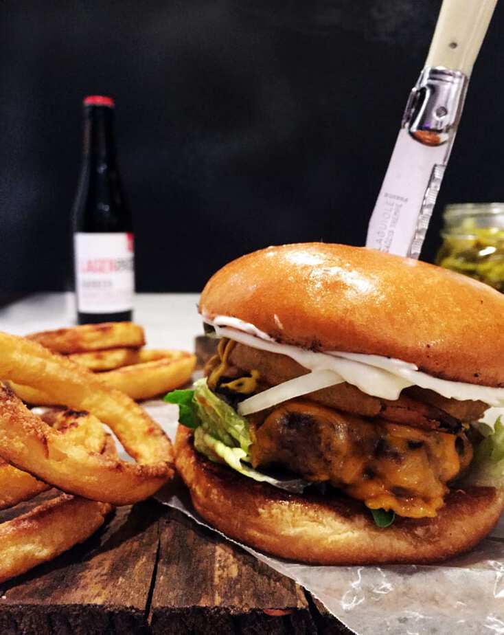 Thick, juicy burger on a glossy egg bun, set on a wood table with a side of golden onion rings beside.