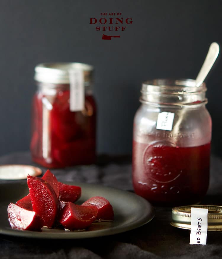 Moody photo with a dark background featuring a black plate filled with dark red pickled beets and mason jars filled with beets.