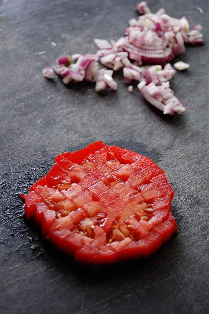 One slice of tomato diced, still in shape of tomato slice with red onion in background on black stone counter.