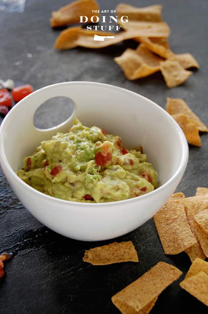 Small white bowl filled with homemade guacamole surrounded by pita chips on a black stone counter.