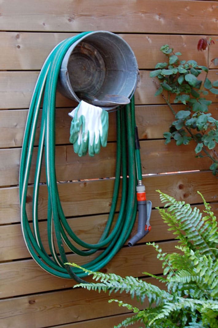 Galvanized bucket screwed to fence with hose wrapped around it and gloves hanging out.