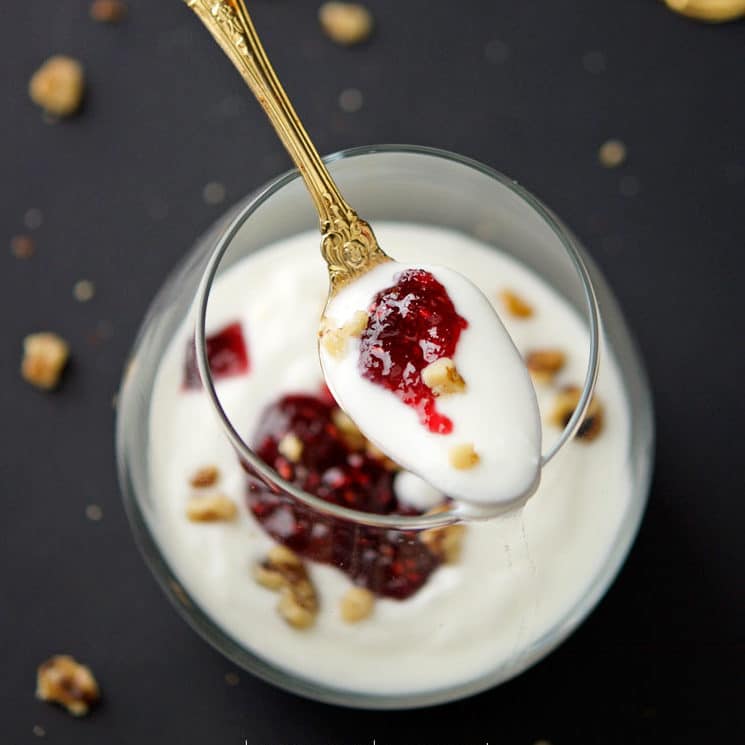 An ornate gold spoon full of yogurt topped with pecans and strawberry jam resting on an elegant glass filled with yogurt.