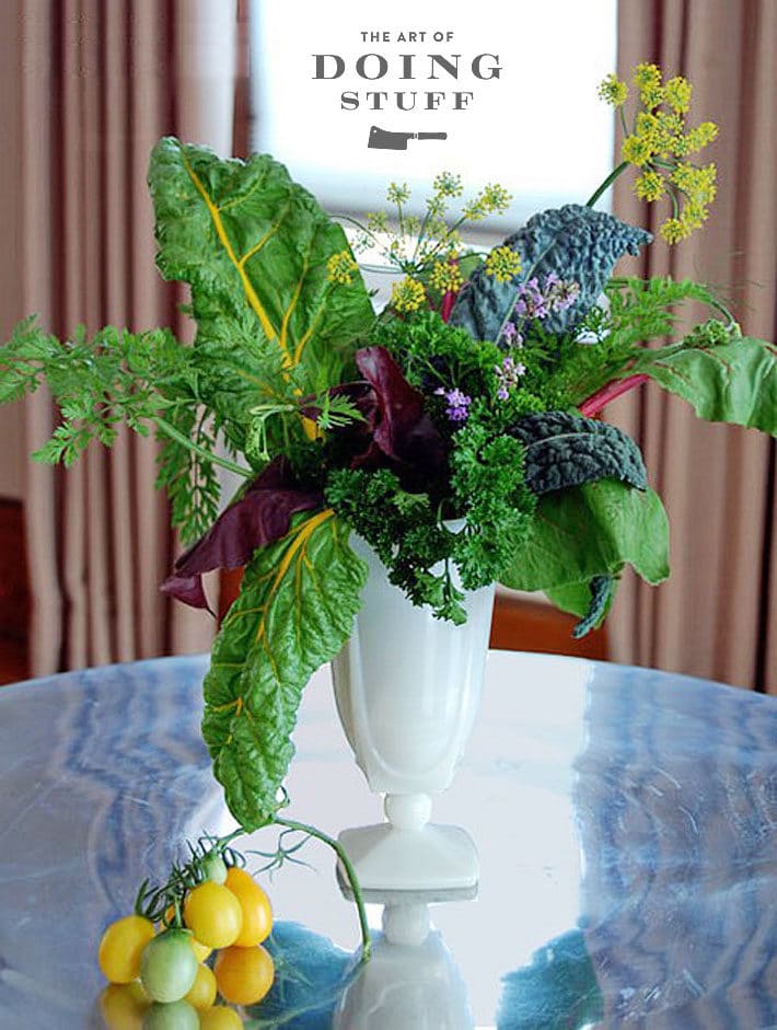 White milkglass vase with an arrangement of vegetables including chard, kale, beet greens, carrot tops and tomatoes.