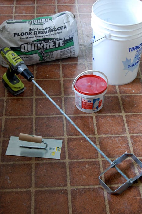 All the materials you need for DIYing self leveling cement laid on the floor including a bucket, self leveling concrete, a drill with cement bit and trowel.
