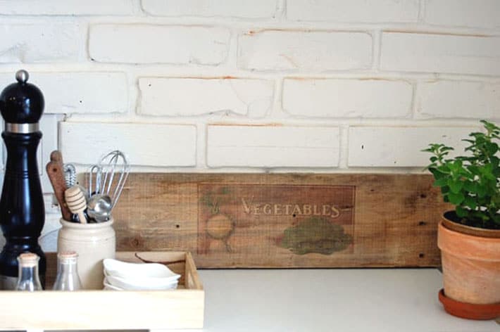 Raw wood with image of vegetables printed on it leaning against white brick wall in farmhouse kitchen.