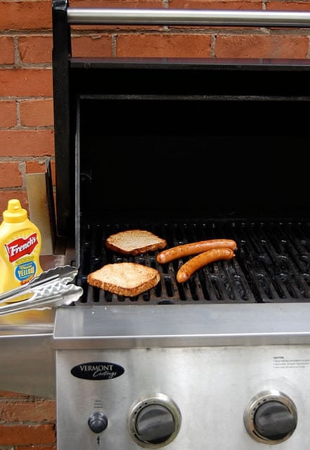 2 hotdogs cooking on a BBQ with a bottle of French's mustard to the side.