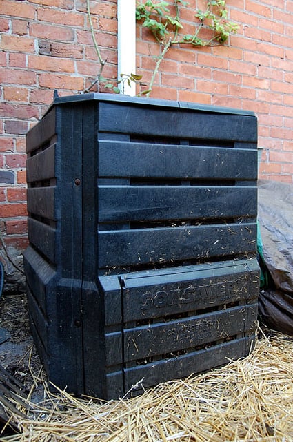 A standard black, square compost bin provided by many municipalities sits against a brick wall.