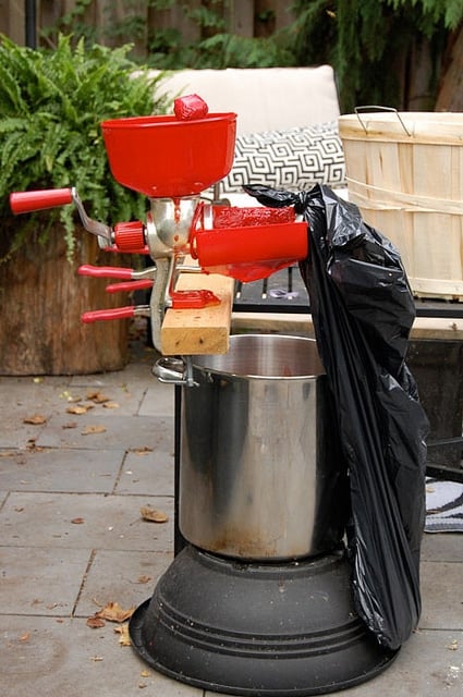 Manual tomato press clamped to a black outdoor table over a stainless steel pot in outdoor courtyard.