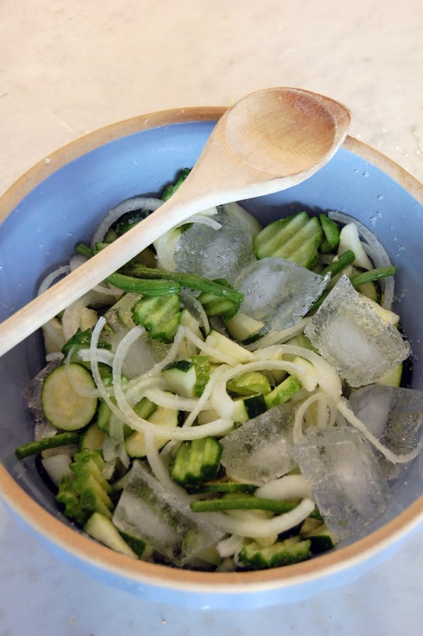 An antique blue bowl filled with salt, bread and butter pickles and ice cubes with a wood spoon laying across the top.