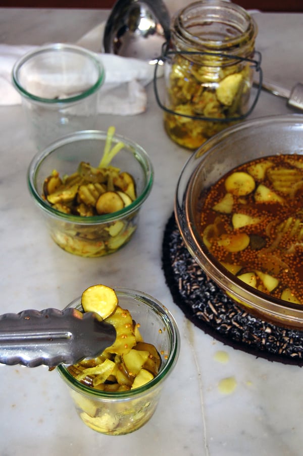 Silver tongs place sliced pickles into a canning jar.