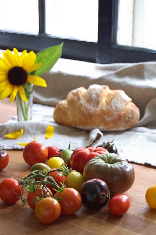 A variety of heirloom tomatoes on a butcher block kitchen counter with bread and a sunflower in the background.