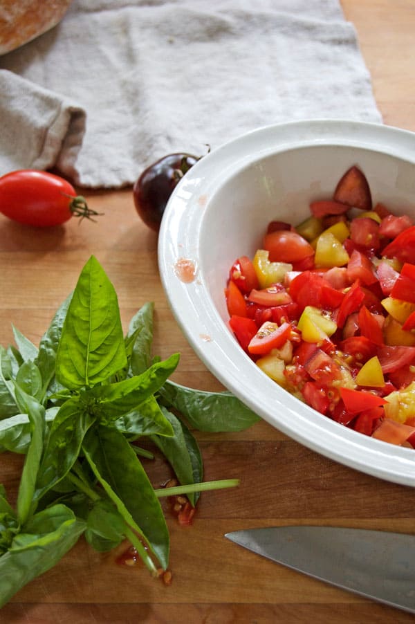 Diced heirloom tomatoes in an ironstone bowl and a bunch of basil sit on a wood countertop.
