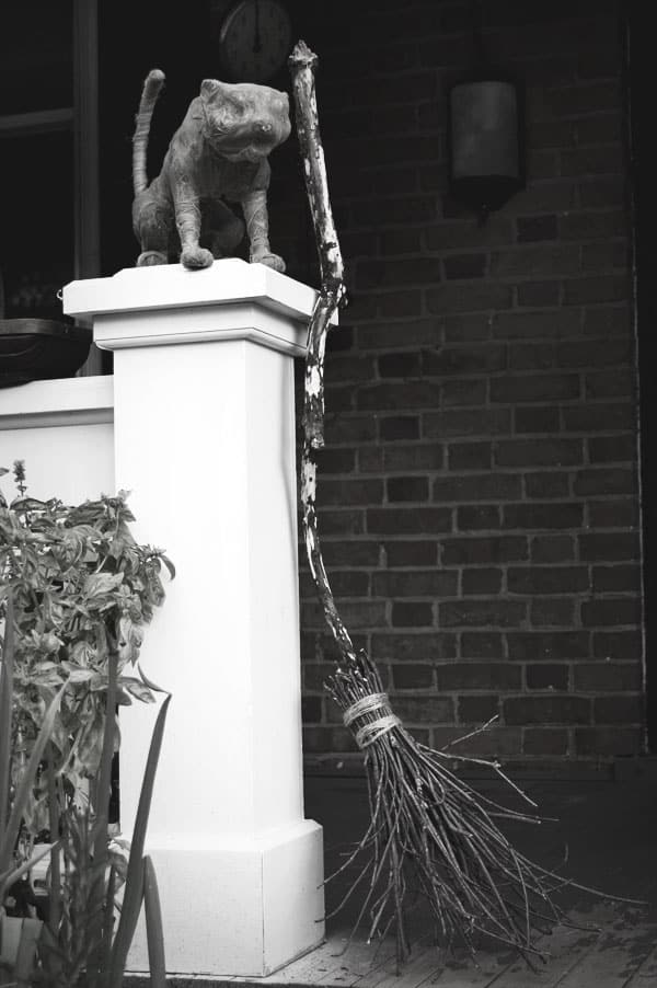 A (fake) mummified cat sits on top of a porch pillar with a witches broom beside it.