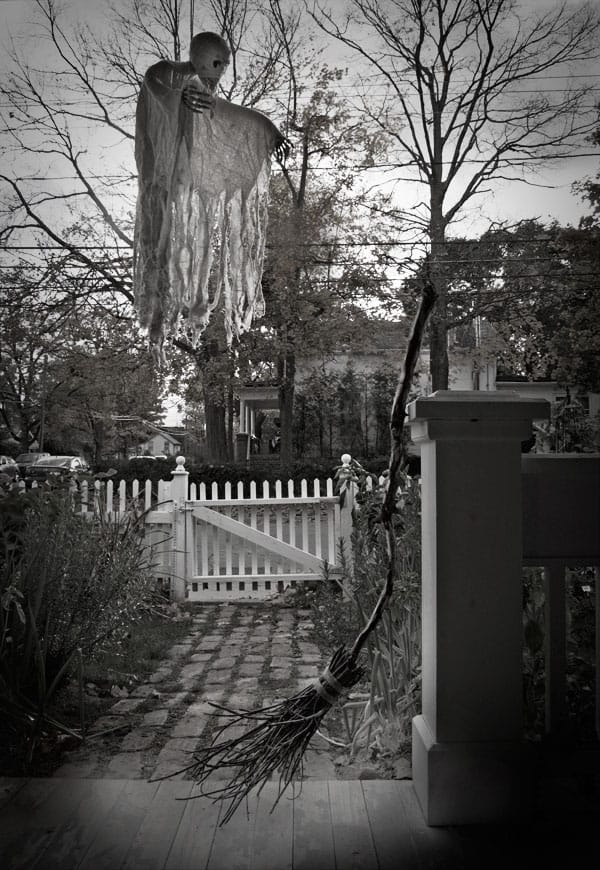 Creepy black and white photo of a witches broom and floating skeleton on a porch with leafless trees in the background.