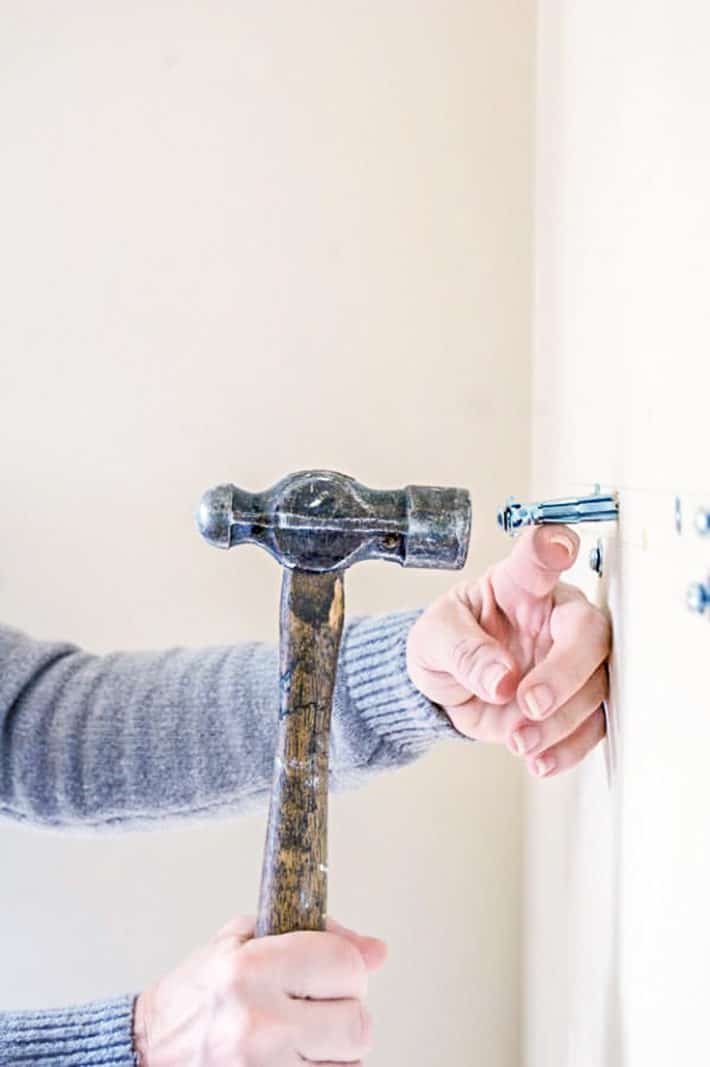 Hammering a hollow wall anchor into a white wall using an old, wood handled hammer.