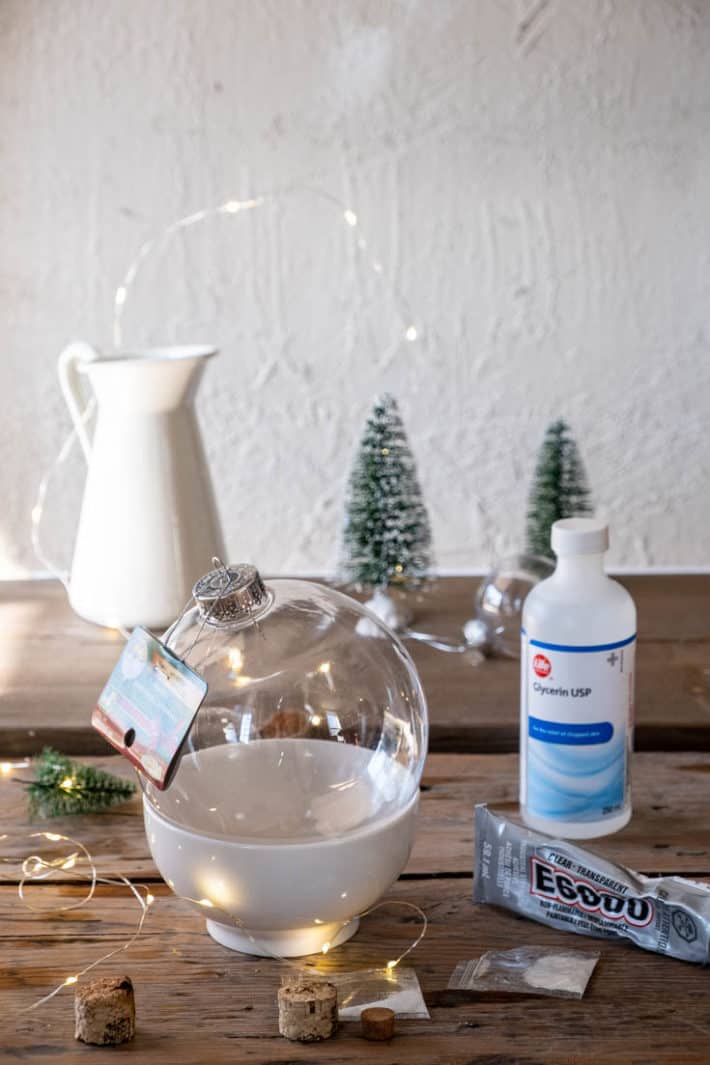 Materials for making a snow globe laid out including a pitcher of water, a clear plastic Christmas ball and tiny bottle brush trees.