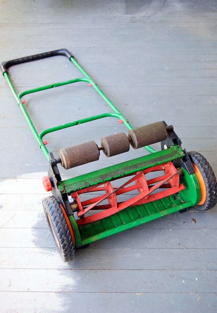 How to sharpen your push mower's blades. Green and red push mower laying down on grey painted porch.