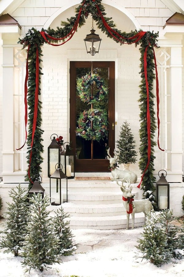 White house showing a door with two wreaths hung vertically, and garland swaged with thin red ribbon over steps surrounded by small evergreen trees.