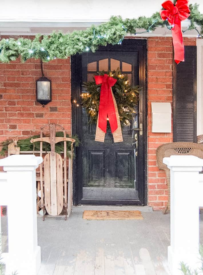 An antique wood sleigh sits beside a black front door on a red brick house decorate with a simple evergreen wreath with a large red bow.