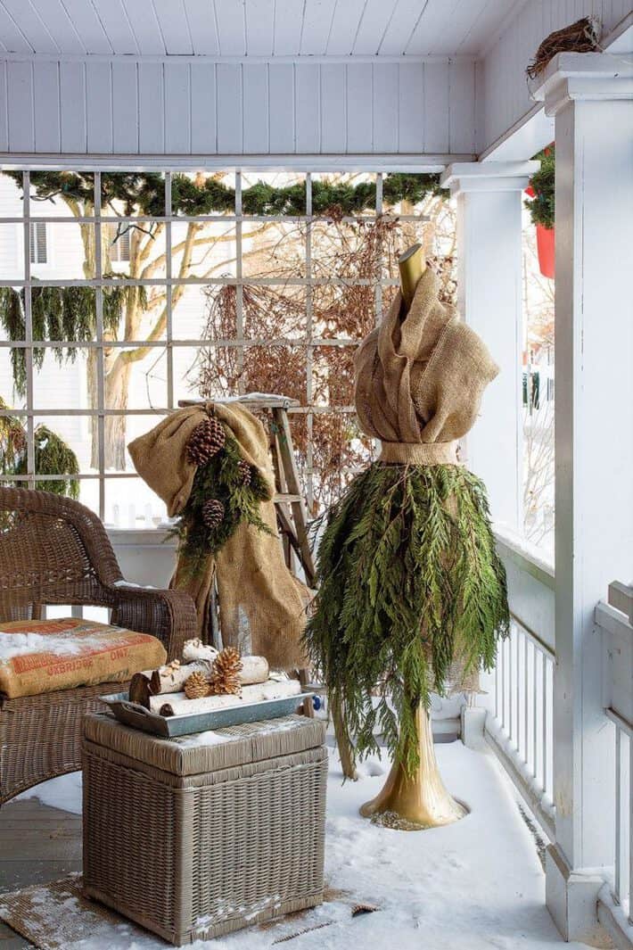 A mannequin wearing a skirt made out of evergreen branches and burlap shaped into a jacket sits on a snow dusted front porch at Christmas.