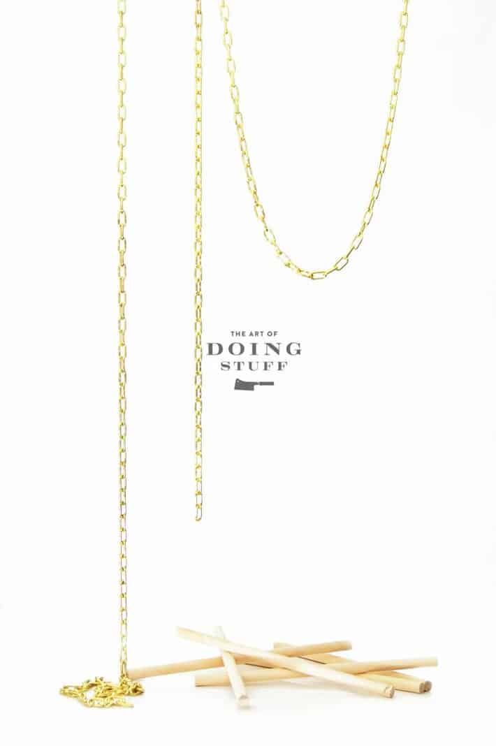 Swaged gold chain hanging from the air on white background with thin wood dowels on ground.