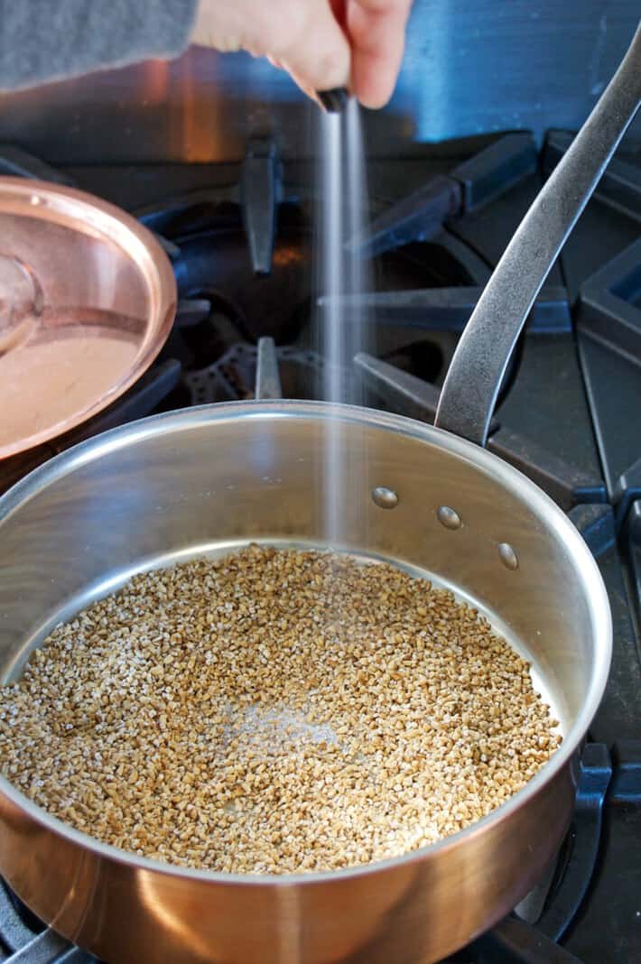 Adding pinch of salt from fingers to oatmeal in copper pot.