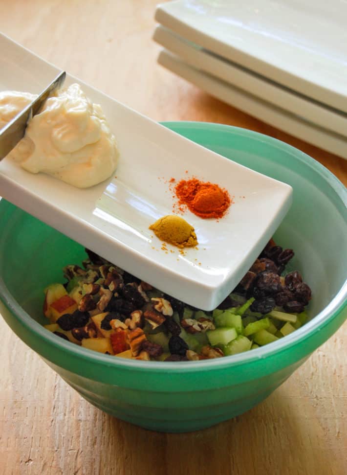 White plate with mayo and spices on it being added to bowl of raisins, celery apples and chicken.