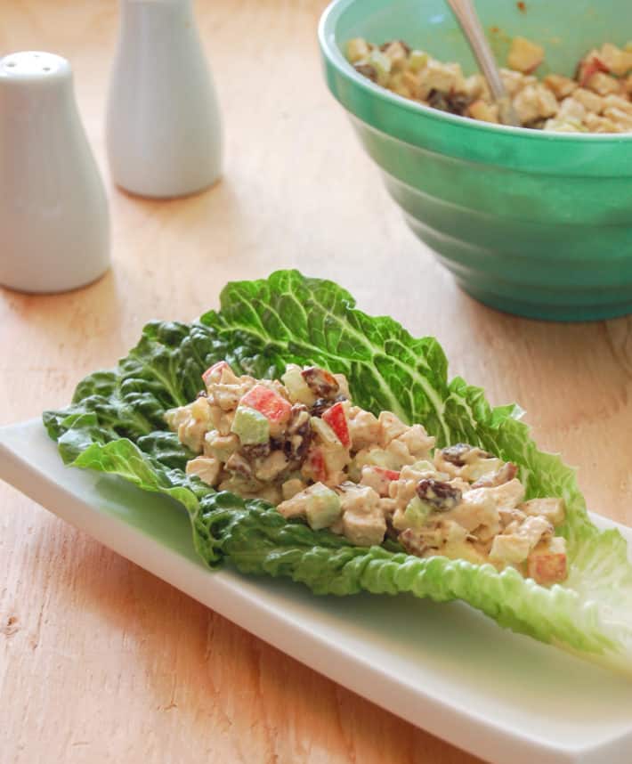 Chicken salad bright with apples celery and raisins, served on a crisp lettuce leaf on a white plate.